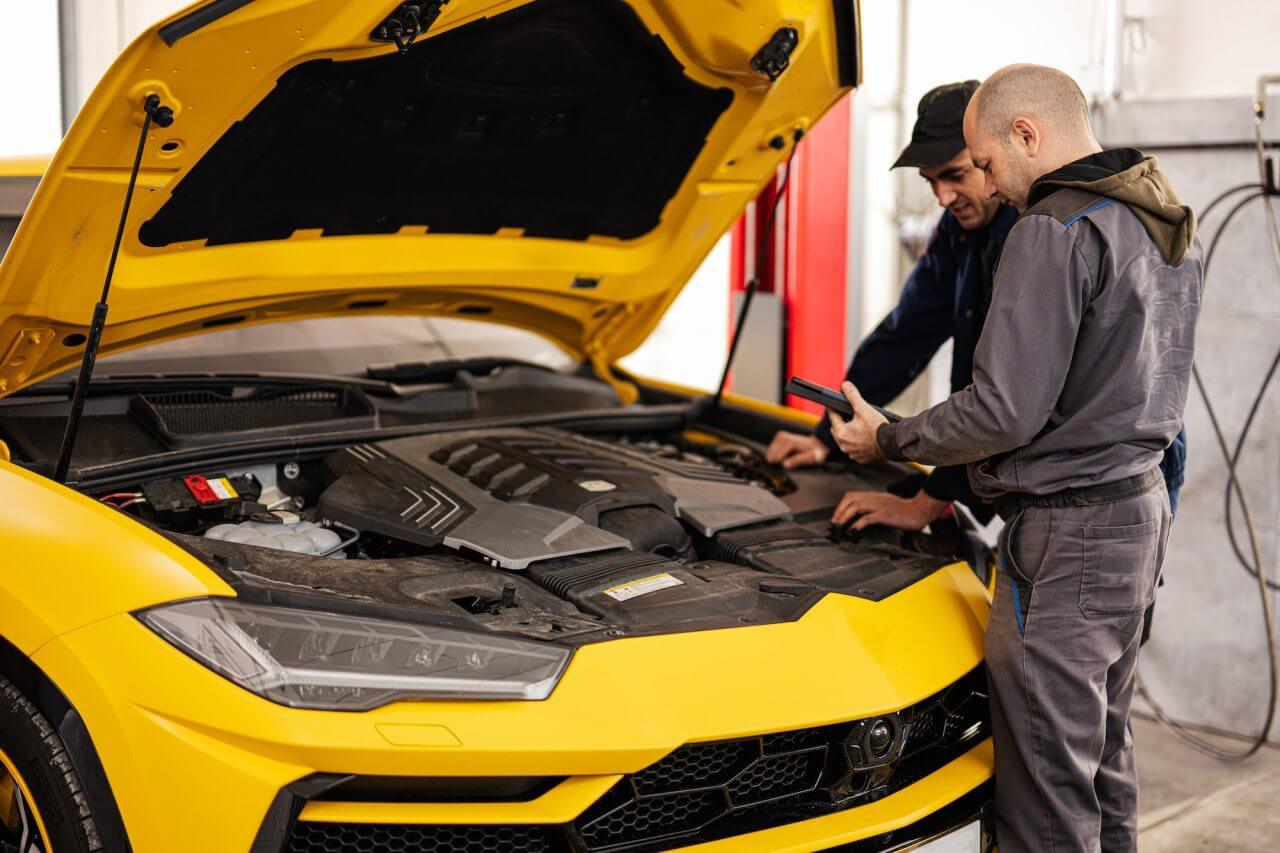 two mechanics stand with tbalet against open hood of yellow sport car suv